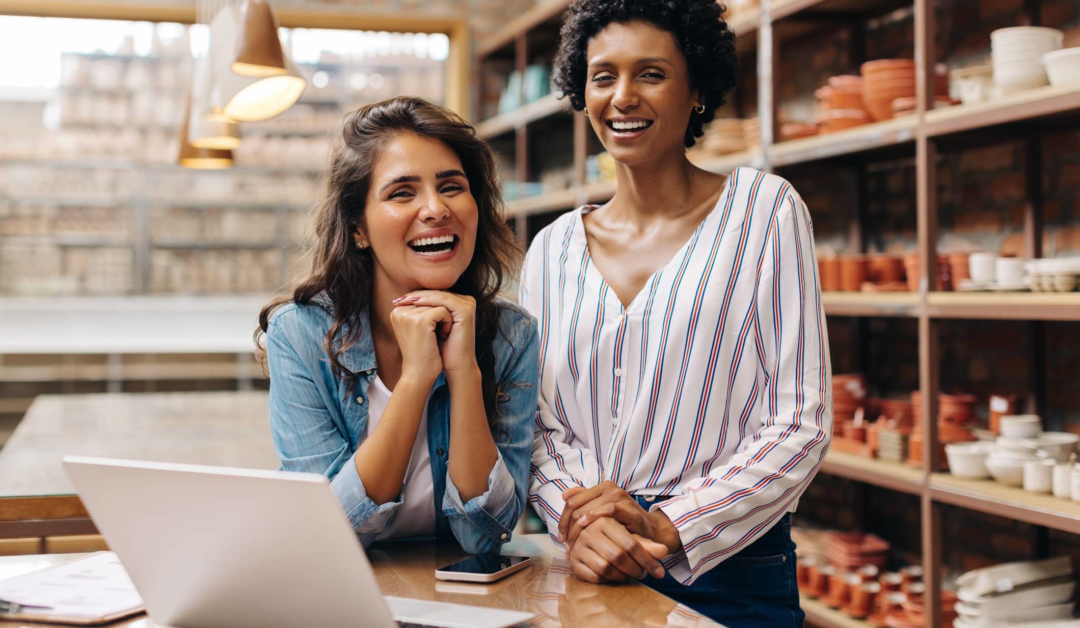 Successful shop owners looking at the camera while working together. Two ceramists using wireless technology in their ceramic store. Happy young businesswomen running a creative small business.